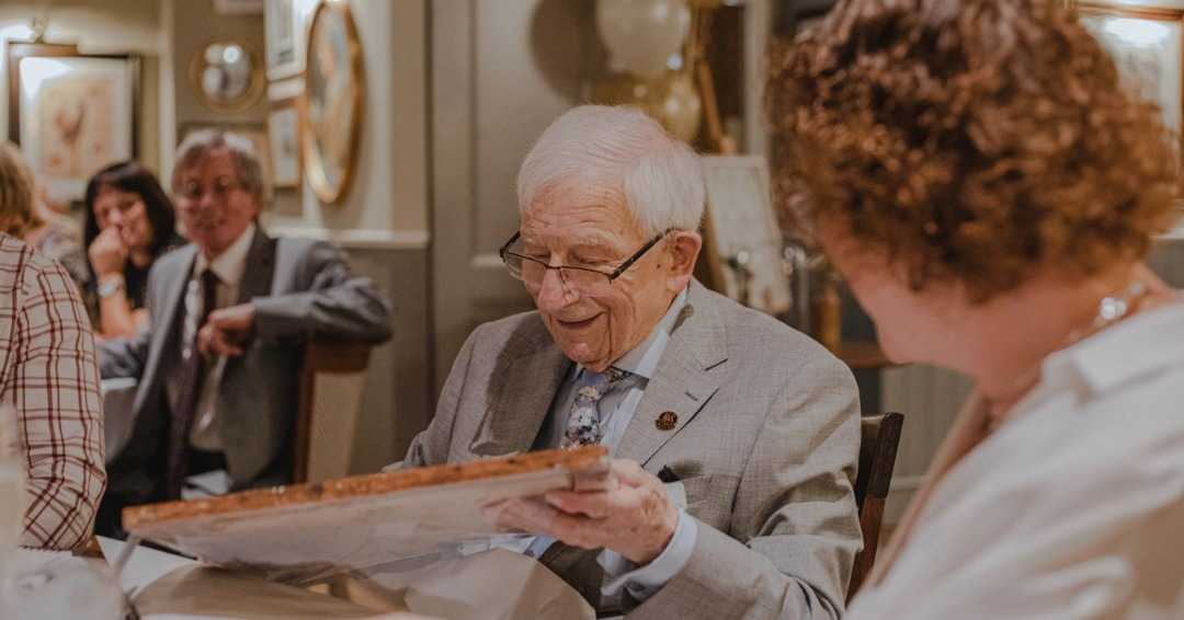an elderly man opening a gift while surrounded by friends