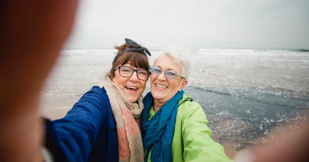 two elderly women with glasses smiling while taking a selfie at the beach